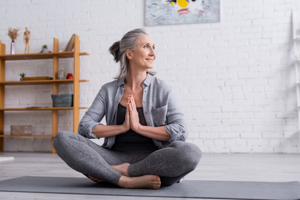 mujer madura feliz con el pelo gris sentado con las manos de oración en la pose de loto en la esterilla de yoga - Foto, Imagen