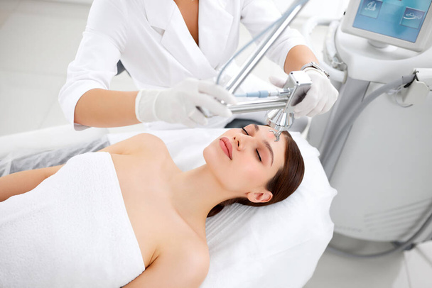 Professzionális kozmetológus lézerberendezéssel bőrápolási kezeléshez a női ügyfél arcán a modern szépségklinikán - Fotó, kép