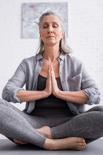 mature woman with praying hands and closed eyes sitting in lotus pose on yoga mat - 写真・画像