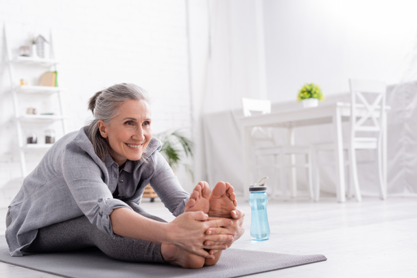 cheerful mature woman with grey hair stretching on yoga mat near sports bottle  - Photo, Image
