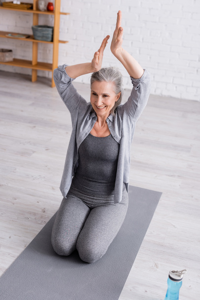 mature woman with praying hands above head practicing yoga near sports bottle  - Photo, Image