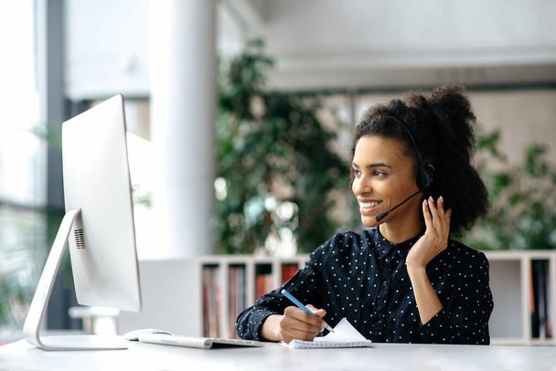 African American friendly young woman with headset, call center worker, consultant, business person, looks at the computer monitor while communicate with colleagues or clients, conducts consultation - Φωτογραφία, εικόνα