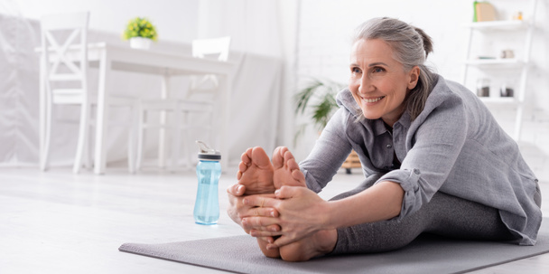 happy mature woman with grey hair stretching on yoga mat near sports bottle, banner - Photo, Image