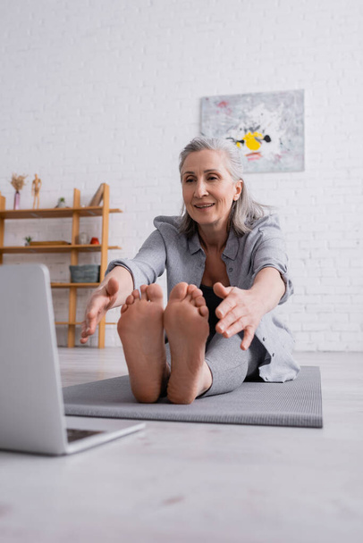 smiling mature woman with grey hair stretching on yoga mat near laptop on blurred foreground  - Zdjęcie, obraz