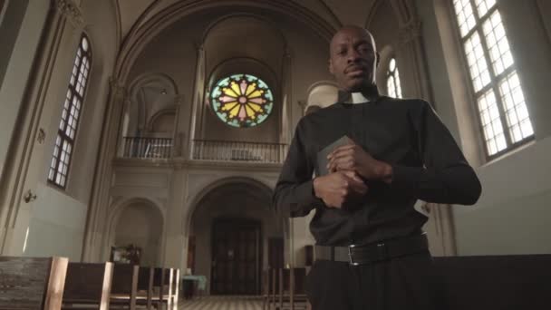 Visão de baixo ângulo do jovem sacerdote afro-americano careca vestindo roupas pretas e colarinho em pé na igreja cristã com a Bíblia nas mãos e olhando para a câmera - Filmagem, Vídeo
