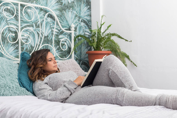 vrouw liggend in bed het lezen van een boek, brunette vrouw in grijze pyjama leest een boek liggend op een bed met een witte deken, naast het bed is er een varen - Foto, afbeelding