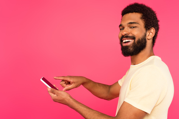 happy african american man pointing at smartphone isolated on pink - 写真・画像