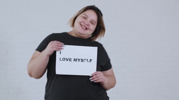 Vücut pozitifliği kavramı - tombul, gülümseyen bir kadın dans ediyor ve üzerinde "Ben de kendimi seviyorum" yazan bir tabela tutuyor. - Video, Çekim