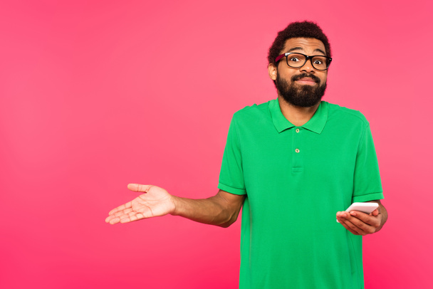hämmentynyt afrikkalainen amerikkalainen mies vihreä t-paita tilalla älypuhelin eristetty vaaleanpunainen - Valokuva, kuva