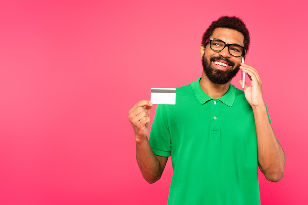 šťastný africký Američan v brýlích mluvit na smartphone a držení kreditní karty izolované na růžové - Fotografie, Obrázek
