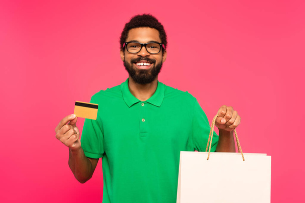 glücklich afrikanisch-amerikanischer Mann in Brille mit Kreditkarte und Einkaufstasche isoliert auf rosa - Foto, Bild