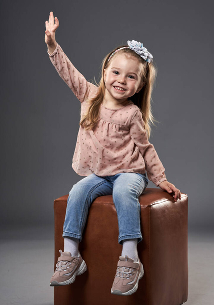 Little adorable girl waving while sitting on a cube chair, isolated on gray background - Valokuva, kuva