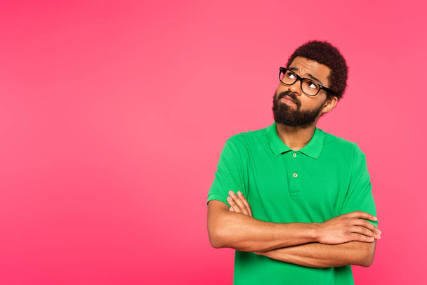 hombre afroamericano pensativo en gafas mirando hacia otro lado mientras está de pie con los brazos cruzados aislados en rosa - Foto, Imagen