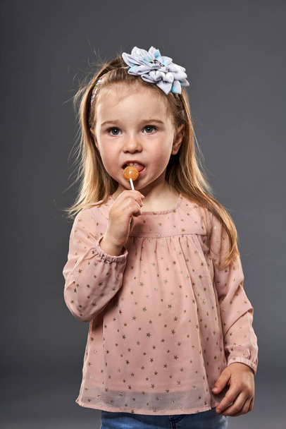 Little blond girl savouring a lollipop, closeup shot isolated on gray background - Фото, изображение