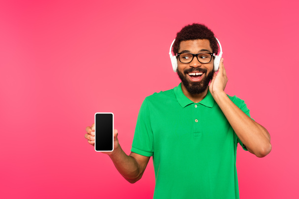 african american man in glasses and wireless headphones holding smartphone with blank screen isolated on pink - Zdjęcie, obraz