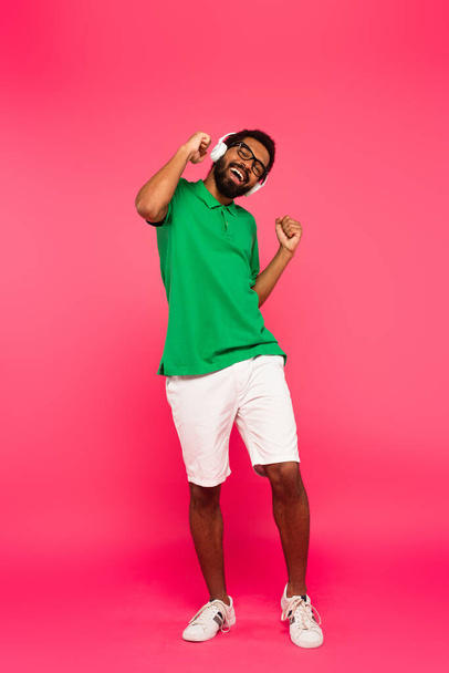 full length of excited african american man in glasses and wireless headphones listening music isolated on pink - Fotó, kép