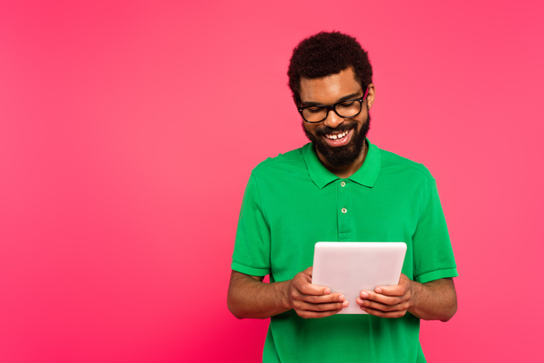 onnellinen afrikkalainen amerikkalainen mies lasit ja vihreä poolopaita käyttäen digitaalinen tabletti eristetty vaaleanpunainen  - Valokuva, kuva