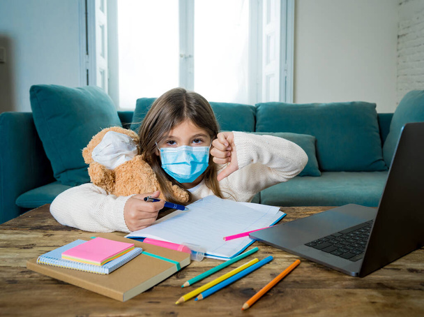 E-learning. Bored and depressed girl with teddy bear and face mask on laptop studying at home in online classes as school remain closed due to New COVID-19 lockdown or forecast weather conditions. - Фото, изображение