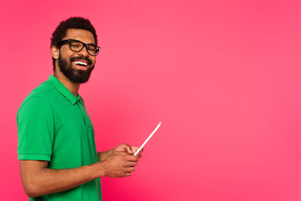wesoły Afroamerykanin w okularach i zielonej koszuli polo za pomocą cyfrowej tabletki odizolowane na różowy  - Zdjęcie, obraz