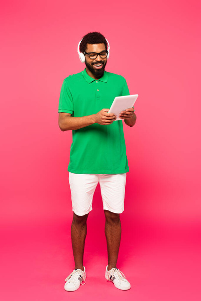 ピンクのデジタルタブレットを使用してメガネやヘッドフォンで陽気なアフリカ系アメリカ人の男の完全な長さ - 写真・画像