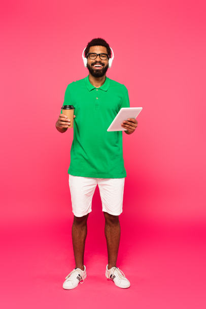 plná délka veselého afrického Američana v brýlích a sluchátkách s papírovým kelímkem a digitálním tabletem na růžové - Fotografie, Obrázek