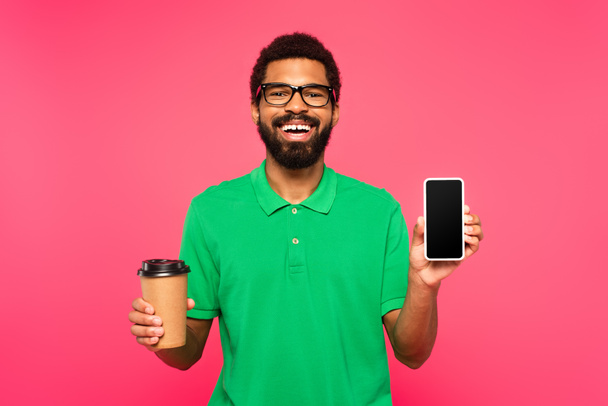 happy african american man in glasses and green polo shirt holding paper cup and smartphone with blank screen isolated on pink - Foto, immagini