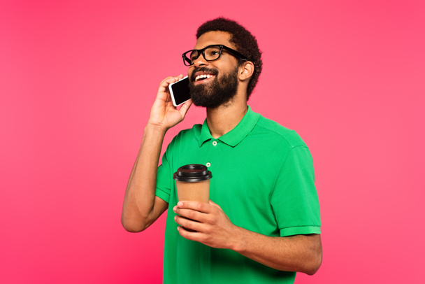 homem americano africano em óculos e camisa polo verde falando no smartphone enquanto segurando copo de papel isolado no rosa - Foto, Imagem