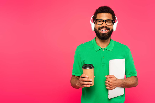 szczęśliwy Afroamerykanin człowiek w okularach i zielonej koszuli polo trzymając papierowy kubek z laptopem izolowane na różowy - Zdjęcie, obraz