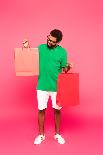 full length of happy african american man in glasses and green polo shirt holding shopping bags on pink - 写真・画像