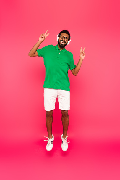 full length of happy african american man in headphones showing peace sign and jumping on pink - Фото, зображення