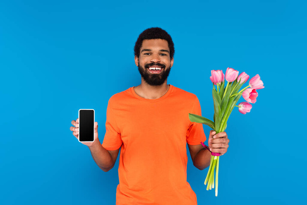 happy african american man holding pink tulips and smartphone isolated on blue - Fotografie, Obrázek