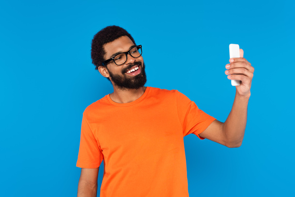 vousatý africký Američan v brýlích s úsměvem a přičemž selfie izolované na modré - Fotografie, Obrázek