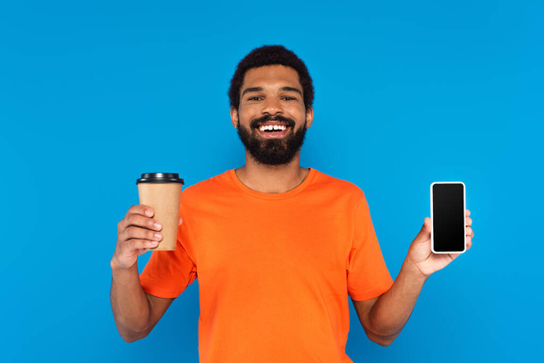 happy african american man holding paper cup and smartphone with blank screen isolated on blue - Zdjęcie, obraz