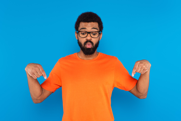 homme afro-américain dans des lunettes regardant vers le bas et pointant avec les doigts isolés sur bleu - Photo, image