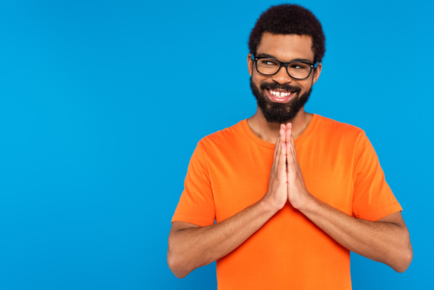 happy african american man in glasses with praying hands isolated on blue - Fotó, kép