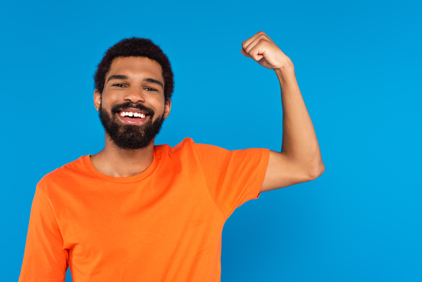 homme afro-américain barbu souriant et montrant le muscle isolé sur bleu - Photo, image
