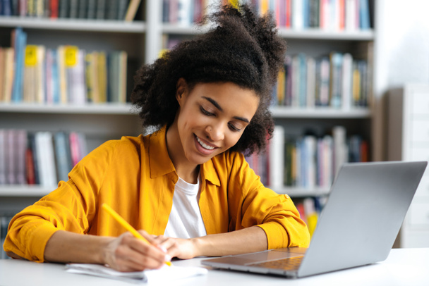 Happy african american smart female student with curly hair, trendy dressed, is sitting at a desk in the university library, with a laptop, focused taking notes during a lecture, smiling - Photo, Image