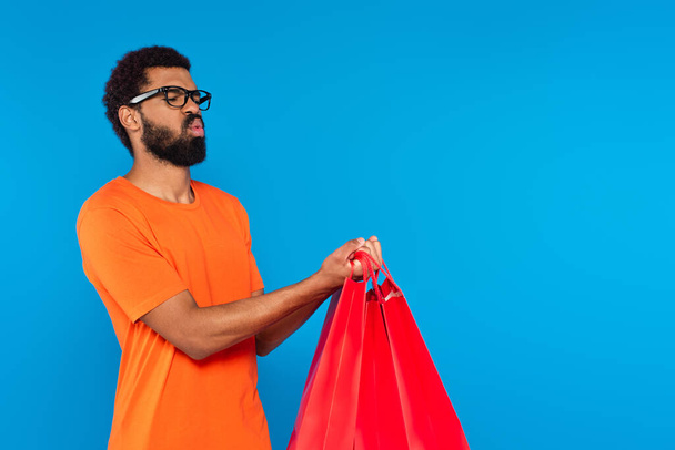 african american man in glasses holding shopping bags and pouting lips isolated on blue  - Photo, Image