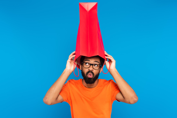 surprised african american man in glasses holding shopping bag above head isolated on blue  - Photo, Image