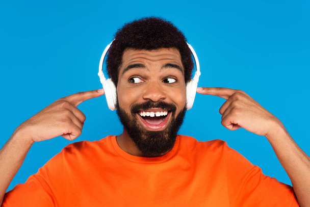 joyful african american man pointing at wireless headphones while listening music isolated on blue - Fotografie, Obrázek