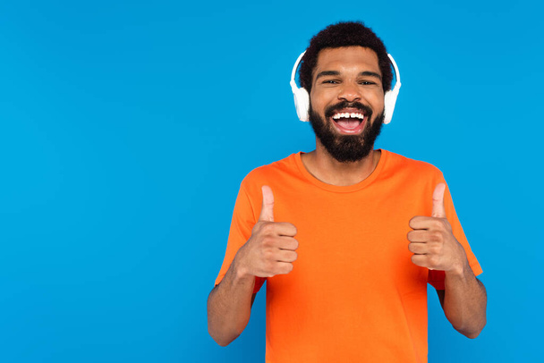 χαρούμενος Αφροαμερικάνος με ασύρματα ακουστικά ακούγοντας μουσική ενώ δείχνει τους αντίχειρες επάνω απομονωμένους στο μπλε - Φωτογραφία, εικόνα