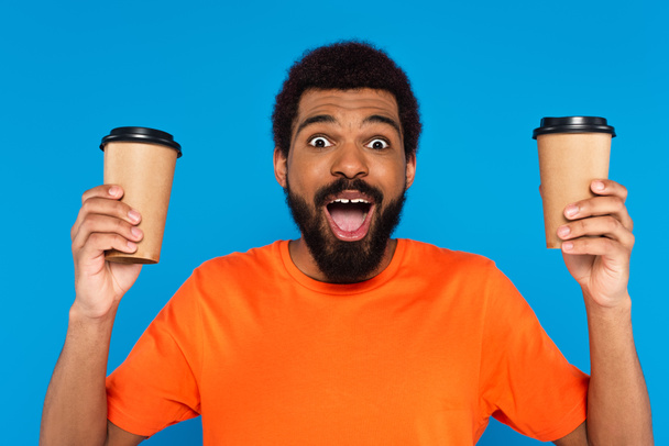 amazed african american man holding paper cups isolated on blue - Foto, Imagen