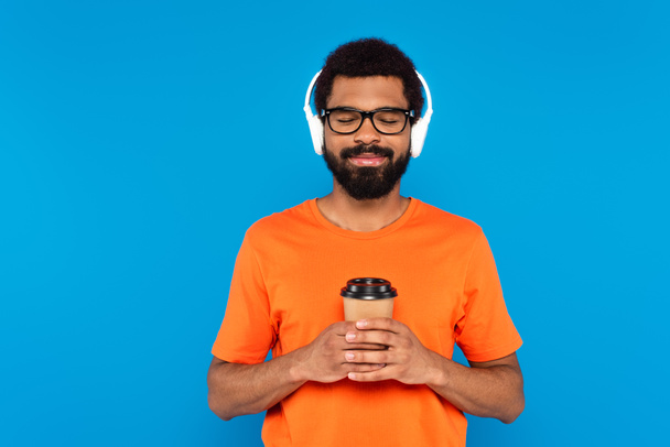 joyful african american man in wireless headphones holding paper cup isolated on blue - Photo, Image