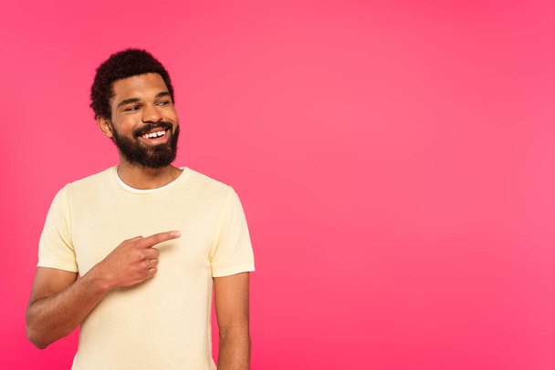cheerful and bearded african american man smiling while pointing with finger isolated on pink  - Фото, зображення