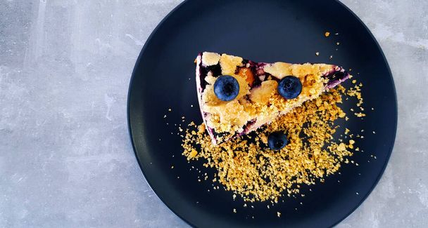 Κάτοψη του Blueberry crumble σε μαύρο πιάτο ή πιάτο σε γκρι φόντο σκυρόδεμα με αντίγραφο χώρο στα αριστερά. Επίπεδη στρώση επιδορπίου στο γκρίζο τραπέζι του καφέ. Χειροποίητο φαγητό με φρούτα και κράκερ. - Φωτογραφία, εικόνα