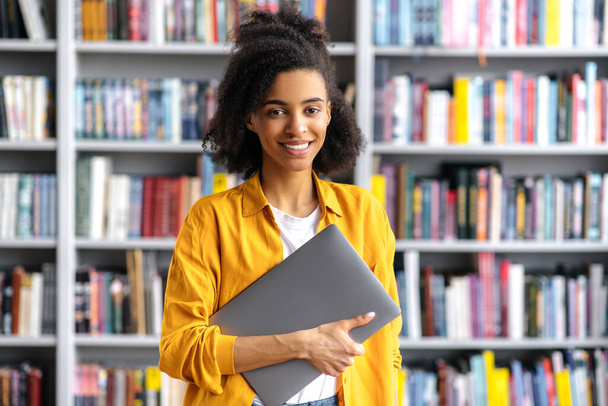 Portret van een studente. Een Afro-Amerikaanse knappe, slimme, zelfverzekerde studente, stijlvol gekleed, met een laptop in de universiteitsbibliotheek, kijkend naar de camera, lachend vriendelijk. - Foto, afbeelding