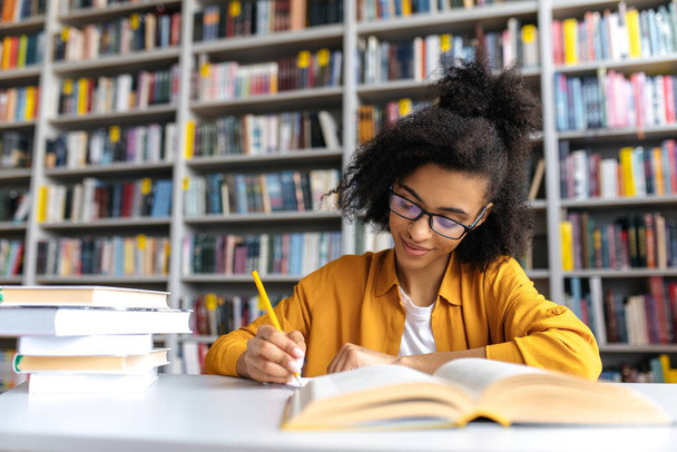 Studentessa afroamericana adolescente che studia mentre siede a tavola nella biblioteca del college, legge libri alla ricerca di informazioni per una lezione o un esame, facendo i compiti e gli appunti, acquisendo conoscenze - Foto, immagini
