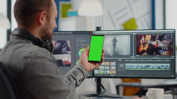 Editor de vídeo hablando en videollamada sosteniendo smartphone con pantalla verde - Metraje, vídeo