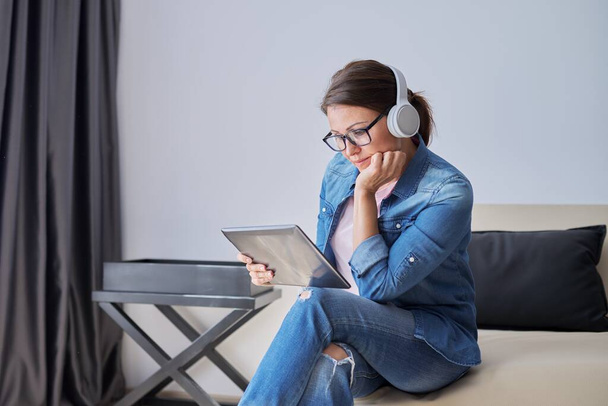 Serious middle aged woman in headphones with digital tablet at home on couch - Photo, Image