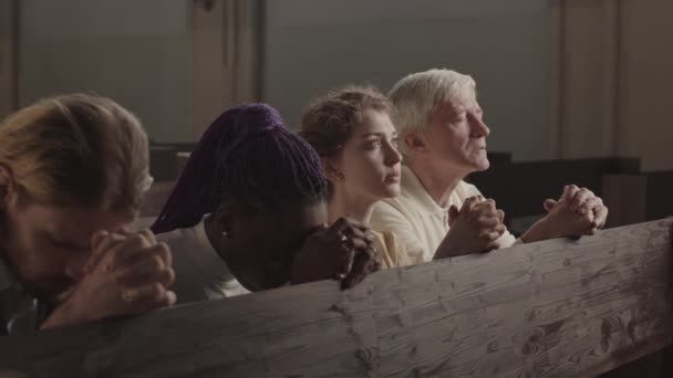 Tracciamento a sinistra di quattro parrocchiani di diversa età e razza seduti su una panchina di legno nella chiesa luterana con gli occhi chiusi e le mani incrociate per la preghiera e la preghiera - Filmati, video